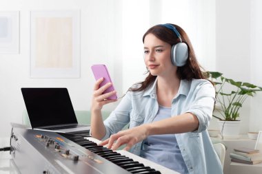 Evde müzik klavyesi çalan, kulaklıkla dinleyen ve akıllı telefonuyla gezinen genç bir kadın. Müzik öğrenmek ve bestelemek. Yaratıcı iş kavramı, müzikle dolu bir yaşam tarzı hayal etmek.