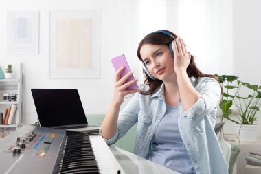 Evde müzik klavyesi çalan, kulaklıkla dinleyen ve akıllı telefonuyla gezinen genç bir kadın. Müzik öğrenmek ve bestelemek. Yaratıcı iş kavramı, müzikle dolu bir yaşam tarzı hayal etmek.