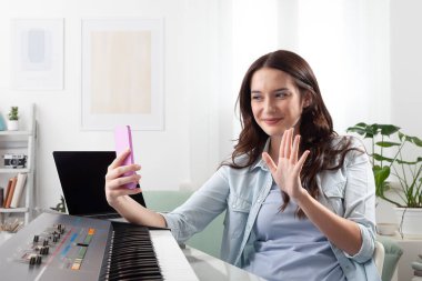 Evde klavye çalan ve arkadaşlarını selamlamak için akıllı telefonunu kullanan genç bir kadın. Çevrimiçi müzik öğrenmek ve bestelemek, uzaktan başkalarıyla işbirliği yapmak. Yaratıcı yaşam tarzı kavramı