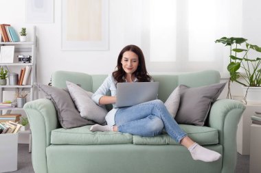 Genç, gülümseyen bir kadın evinde rahat rahat kanepede oturuyor, parlak bir oturma odasında dizüstü bilgisayarla çalışıyor. Günlük yaşam tarzı, uzaktan çalışma, internette gezinme, rahat ev ortamı