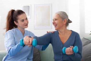 Bir huzurevinde fizik tedavi seansı sırasında hafif halterli yaşlı bir kadına yardım eden gülümseyen genç hemşire. Kıdemli hasta mutlu ve meşgul görünüyor. Sağlık hizmetlerinden destek alıyor..
