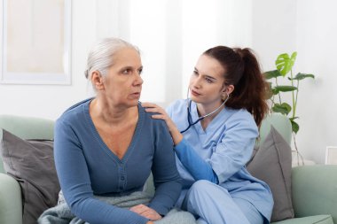 Gülümseyen genç hemşire ya da doktor, klinikte ya da evde steteskoplu yaşlı bir kadının kalp atışlarını ve nefes alışlarını dinliyor. Sağlık kontrolü ve sağlık hizmeti kavramı.