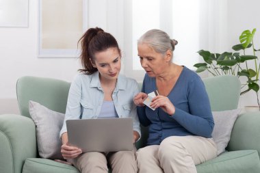 Torunum, kanepede oturan büyükannem, yaşlı kadın ve genç bayana dizüstü bilgisayar ve kredi kartıyla online alışveriş yapmalarında yardım ediyor. Aile kavramı, teknoloji ve dijital destek