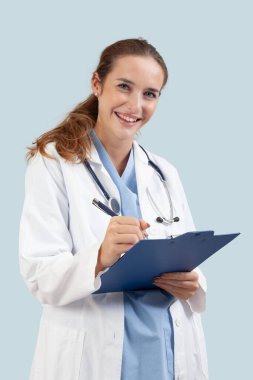 Beyaz önlüklü gülümseyen kadın doktor bir reçete ya da hasta notlarını panoya yazıyor. Steteskopu açık mavi arka plan, sağlık ve tıbbi konsept üzerine izole edilmiş tıbbi uzman..