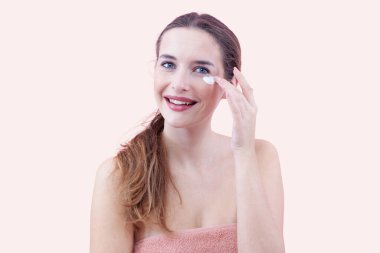 Gülümseyen genç bir kadın kameraya bakarken cilt bakım nemlendiricisi sürüyor. Cilt bakımı ve güzellik konsepti. Pembe arka planda izole edilmiş. Yumuşak, makyajlı ve sağlıklı ciltler için yüz bakımı