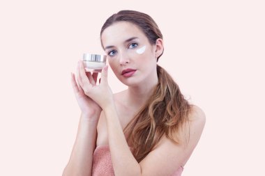 Cilt nemlendirici süren ve kameraya bakarken bir kavanoz krem tutan genç bir kadın. Cilt bakımı ve güzellik anlayışı. Pembe arka planda izole edilmiş. Yüz bakımı, makyaj ve sağlıklı cilt