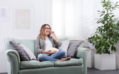 Akıllı gözlük takan, elinde patlamış mısır tutan ve gözlüklerine dokunan bir kadın sanki sanal bir ekranla etkileşim içindeymiş gibi. Evdeki dijital eğlencenin tadını çıkarıyorum. Gelecekçi teknoloji konsepti