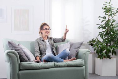 Akıllı gözlüklü gülümseyen genç bir kadın, evinde yanında akıllı telefonuyla koltukta oturuyor. İleriye bakıyor ve ekrana dokunuyor, dijital içerikle etkileşime geçiyor. Gelecekçi teknoloji