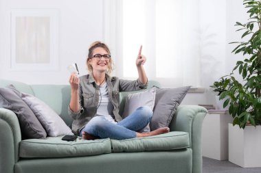 Akıllı gözlükler takan genç bir kadın, evinde yanında akıllı telefonuyla bir koltukta oturuyor, dokunmatik ekranda kredi kartı tutuyor. Dijital ödeme ve online alışveriş, teknoloji yaşam tarzı