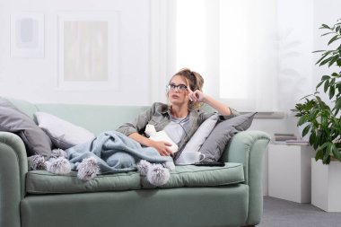 Acı çeken bir kadın, evinde bir kanepede yatarken karnına sıcak su şişesi tutarken, akıllı gözlükler takıyor ve tıbbi yardım veya dijital sağlık hizmetleri çözümleri bulmak için sanal ekranla etkileşime geçiyor.