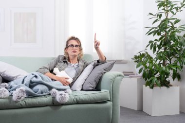 Acı çeken bir kadın, evinde bir kanepede yatarken karnına sıcak su şişesi tutarken, akıllı gözlükler takıp tıbbi yardım veya dijital sağlık hizmetleri için sanal bir ekrana dokunuyor.