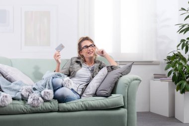 Kanepede uzanmış gülümseyen bir kadın, akıllı gözlükler takıyor ve sanal ekranla iletişim kurarken elinde bir kredi kartı tutuyor. Çevrimiçi alışveriş kavramı, dijital ödeme, evdeki akıllı teknoloji