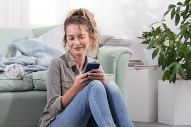 Yerde oturan gülümseyen genç bir kadın, akıllı telefonuyla meşgul. Modern ev ortamında dijital yaşam tarzı, sosyal medya ve çevrimiçi içeriğin tadını çıkarmak.