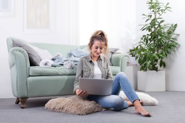 Dizüstü bilgisayarıyla meşgul bir şekilde yerde bağdaş kurmuş gülümseyen genç bir kadın. Modern ev ortamında dijital yaşam tarzı, sosyal medya ve çevrimiçi içeriğin tadını çıkarmak.