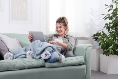 Genç bir kadın kanepeye uzanmış, elinde battaniyeyle dizüstü bilgisayar kullanırken gülümsüyor. Rahat bir ev ortamında dijital yaşam tarzı, sosyal medya ve çevrimiçi içeriğin tadını çıkarmak