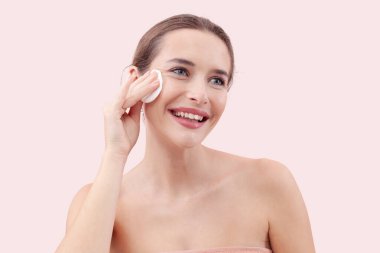 Gülümseyen genç bir kadın yüzünü pamuk ped ve mikiler suyuyla temizliyor. Cilt rutini, makyaj çıkarma, taze sağlıklı deri. Pembe arka plan, güzellik ve hijyen kavramına izole edilmiş.