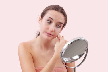 Genç bir kadın, aynanın önünde yüzünü pamuk pedlerle temizliyor ve suyu bodruma indiriyor. Cilt rutini, makyaj çıkarma, taze sağlıklı deri. Pembe arka plan, güzellik ve hijyen kavramına izole edilmiş.