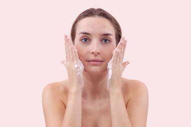 Yüzünü köpük temizleyiciyle temizleyen, ellerini köpükle kaplayan ve cildine süren genç bir kadın. Temiz ve sağlıklı yüz için cilt rutini. Pembe arka planda izole edilmiş, sivilceli.