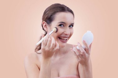 Gülümseyen genç bir kadın makyaj fırçasıyla allık sürüyor, bir ayna tutuyor ve kameraya bakıyor. Temiz, asgari güzellik konsepti. Bej arkaplanda izole edilmiş.