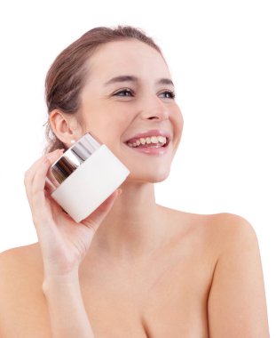 Makyajlı gülümseyen genç bir kadın yan bakarken yüzünde plastik bir yüz kremi kavanozu tutuyor. Cilt bakımı ve güzellik tedavisi rutin konsepti. Beyaz arkaplanda izole