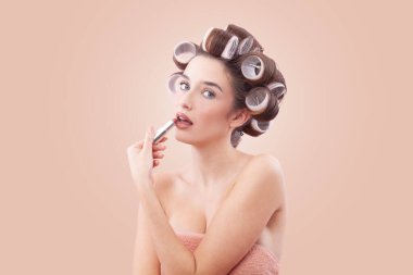 Makyaj, mavi gözleriyle kameraya bakarken saç bigudiyle ruj süren gülümseyen genç kadın. Saç stili, makyaj, güzellik ve kozmetik anlayışı. Bej rengi ile izole edilmiş.