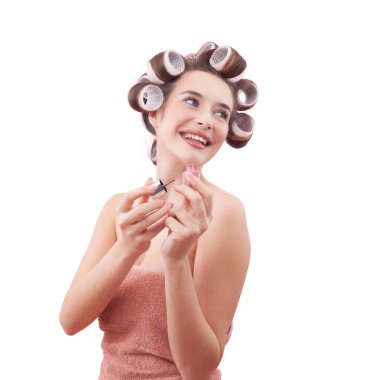 Mavi gözleriyle tırnak cilası sürerken saç bigudiyle gülümseyen genç bir kadın. Saç stili, makyaj, güzellik ve kozmetik anlayışı. Beyaz arkaplanda izole