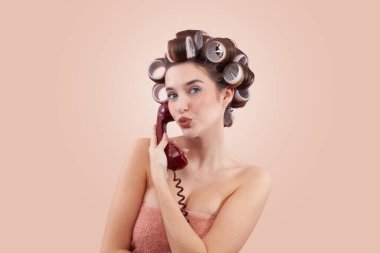 Saç bigudileri olan bir kadın kameraya öpücük gönderirken antika bir telefon kullanıyor. Saç stili, makyaj, güzellik ve kozmetik anlayışı. Muhtemelen kuaför randevusu alıyorum. Bej rengi ile izole edilmiş.