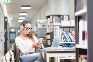 Genç bir üniversite öğrencisi kütüphanede okuyor. Masada oturuyor, kulaklık takıyor ve akıllı telefon kullanıyor. Eğitim, bilgi, modern öğrenme ve sınav hazırlığı kavramı