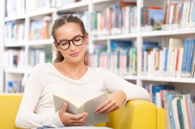 Genç bir üniversite öğrencisi kütüphanede okuyor. O bir koltukta oturuyor ve bir kitap okuyor. Eğitim, öğrenim, akademik başarı ve sınav hazırlığı kavramı.