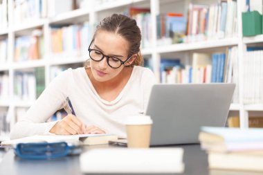 Genç bir üniversite öğrencisi kütüphanede okuyor, bir masada bilgisayarla oturuyor ve bir deftere yazı yazıyor. Eğitim, bilgi, öğrenme, akademik başarı ve sınav hazırlığı kavramı.