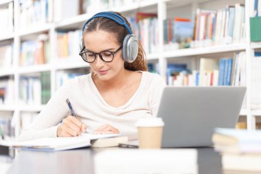 Kütüphanede okuyan genç bir kız öğrenci, bilgisayarlı bir masada oturuyor, kulaklık takıyor ve bir deftere not alıyor. Eğitim, e-öğrenim ve çevrimiçi akademik öğrenim kavramı.