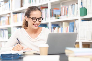 Genç bir üniversite öğrencisi kütüphanede okuyor, bir masada bilgisayarla oturuyor ve bir deftere yazı yazıyor. Eğitim, bilgi, öğrenme, akademik başarı ve sınav hazırlığı kavramı.