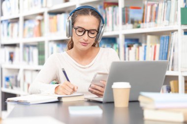 Genç bir üniversite öğrencisi kütüphanede okuyor, bilgisayarlı bir masada oturuyor, kulaklık takıyor, akıllı telefon kullanıyor ve bir not defterine not alıyor. Eğitim kavramı, çevrimiçi öğrenim.