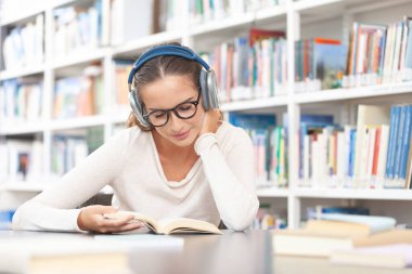 Genç bir üniversite öğrencisi kütüphanede okuyor. Masada oturuyor, kulaklık takıyor ve kitap okuyor. Eğitim, bilgi, öğrenme, akademik başarı ve sınav hazırlığı kavramı.