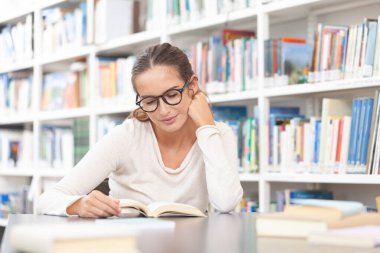 Genç bir üniversite öğrencisi kütüphanede okuyor. O bir masada oturuyor ve bir kitap okuyor. Eğitim, bilgi, öğrenme, akademik başarı ve sınav hazırlığı kavramı.