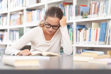 Genç bir üniversite öğrencisi kütüphanede okuyor. O bir masada oturuyor ve bir kitap okuyor. Eğitim, bilgi, öğrenme, akademik başarı ve sınav hazırlığı kavramı.