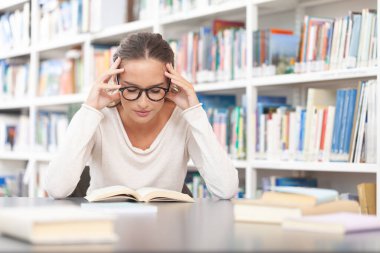 Genç bir üniversite öğrencisi kütüphanede okuyor. O bir masada oturuyor ve bir kitap okuyor. Eğitim, bilgi, öğrenme, akademik başarı ve sınav hazırlığı kavramı.