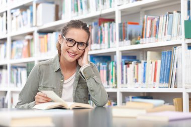 Kütüphanede okuyan gülümseyen genç bir üniversite öğrencisi. O bir masada oturuyor ve bir kitap okuyor. Eğitim, bilgi, öğrenme, akademik başarı ve sınav hazırlığı kavramı.