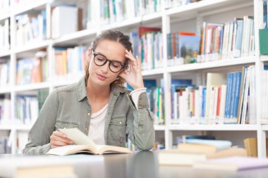 Genç bir üniversite öğrencisi kütüphanede okuyor. O bir masada oturuyor ve bir kitap okuyor. Eğitim, bilgi, öğrenme, akademik başarı ve sınav hazırlığı kavramı.