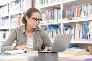 Genç bir üniversite öğrencisi kütüphanede okuyor, bir masada bilgisayarla oturuyor ve bir deftere yazı yazıyor. Eğitim, bilgi, öğrenme, akademik başarı ve sınav hazırlığı kavramı.