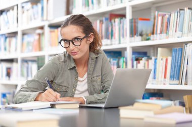 Genç bir üniversite öğrencisi kütüphanede okuyor, bir masada bilgisayarla oturuyor ve bir deftere yazı yazıyor. Eğitim, bilgi, öğrenme, akademik başarı ve sınav hazırlığı kavramı.