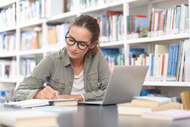Genç bir üniversite öğrencisi kütüphanede okuyor, bir masada bilgisayarla oturuyor ve bir deftere yazı yazıyor. Eğitim, bilgi, öğrenme, akademik başarı ve sınav hazırlığı kavramı.