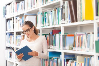 Modern bir kütüphanede kitap okuyan gözlüklü genç bir kadın. Arkaplanda renkli kitaplarla dolu kitaplıklar. Öğrenim, eğitim, öğrenim, bilgi ve akademik yaşam tarzı kavramı