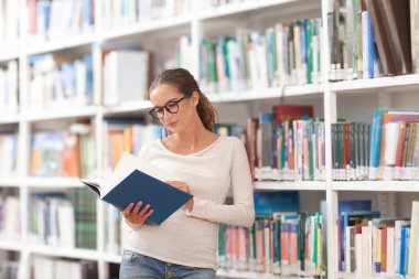 Modern bir kütüphanede kitap okuyan gözlüklü genç bir kadın. Arkaplanda renkli kitaplarla dolu kitaplıklar. Öğrenim, eğitim, öğrenim, bilgi ve akademik yaşam tarzı kavramı