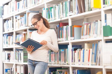 Modern bir kütüphanede kitap okuyan gözlüklü genç bir kadın. Arkaplanda renkli kitaplarla dolu kitaplıklar. Öğrenim, eğitim, öğrenim, bilgi ve akademik yaşam tarzı kavramı