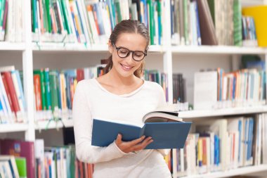 Modern bir kütüphanede kitap okuyan gözlüklü genç bir kadın. Arkaplanda renkli kitaplarla dolu kitaplıklar. Öğrenim, eğitim, öğrenim, bilgi ve akademik yaşam tarzı kavramı