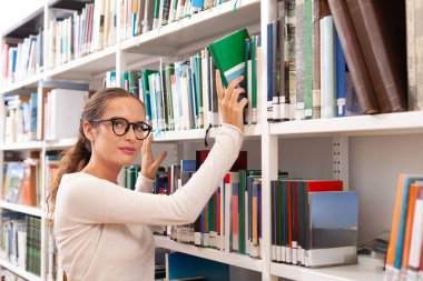 Modern bir kütüphanenin rafından kitap seçen gözlüklü genç bir kadın. Eğitim, bilgi, öğrenme ve akademik araştırma kavramı. Çalışmayı, merakı ve entelektüel yaşam tarzını temsil eder