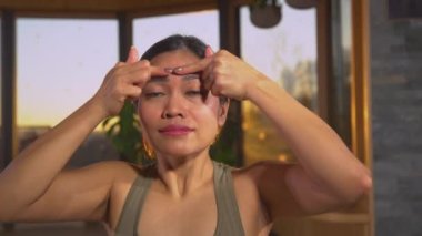 Güzel Asyalı kadın kendini gençleştirme yüz masajı ile tedavi ediyor. Yüz kaslarını gevşetmek için yüz yoga pratiği. Güzel bir kadın, altın ışıkta yüzüne dokunup masaj yapıyor.
