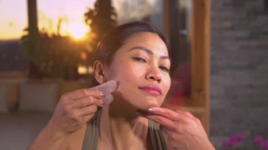 Güzel bir Asyalı kadın yüzüne gua sha Jade kazıyıcısıyla masaj yapıyor. Yenilenme için yüz tedavisi uygulaması. Yaşlanma karşıtı terapinin bir parçası olarak güzel bir kadın yüz masajı yapıyor..