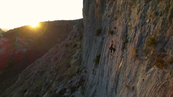 Aerial 背景に金色の太陽のフレアを持つ石灰岩の壁の真ん中に登る女性 日没時に女性登山家が上昇する 美しい自然環境の中でアドレナリン屋外活動 — ストック動画